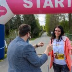 Na zdjęciu kobieta z numerem startowym 198 kończy bieg w parku. Jest uśmiechnięta i trzyma kijki do nordic walking. W tle znajduje się łuk startowy i zalesiona ścieżka.