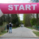 Zdjęcie przedstawia start biegu pod różowo-białym łukiem z napisem START. W tle widoczny jest las i kilka osób na drodze. Po prawej stronie znajduje się wyświetlacz z czasem.