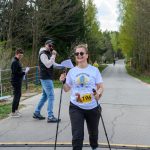 Na zdjęciu widoczna jest kobieta w białej koszulce i okularach, korzystająca z kijków do nordic walking. W tle znajdują się drzewa i dwie osoby z mikrofonem i kartkami. Nad nią rozciąga się różowy łuk, na drodze asfaltowej.