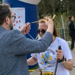 Na zdjęciu mężczyzna zakłada medal uśmiechniętej kobiecie w białej koszulce. Tło to zieleń i ludzie w casualowym ubraniu. Obraz jest pełen radości i celebracji.