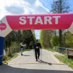 Zawodnik z kijkami przechodzi pod różowo-białym łukiem startowym. Człowiek w tle notuje wyniki. Tło to zielone drzewa i niebo.