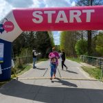 Na zdjęciu osoba w kolorowym stroju przechodzi przez linię startu biegu w lesie. Nad nią dmuchany łuk w kolorach różowym i niebieskim z napisem START. Obok widoczny jest zegar z czasem, a w tle drzewa i droga.