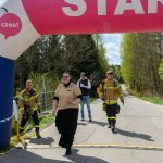 Na zdjęciu jest start biegu w plenerze z nadmuchiwanym łukiem w kolorach różowym i niebieskim. Strażacy idący po asfalcie, otoczeni są zielenią drzew. Obok łuku widoczna jest mapa Polski z zaznaczonymi miejscami.