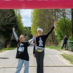 Dwie uśmiechnięte dziewczyny stoją na drodze pod różowym balonem START. Trzymają medale, wokół zieleń lasu. Słoneczny dzień.