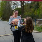 Zdjęcie przedstawia kobietę finiszerkę w biegu z numerem 66. Otrzymuje medale przy linii startu/meta. Tło to drzewa i droga.