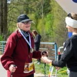 Na zdjęciu dwie osoby rozmawiają na zewnątrz. Osoba po lewej ma czerwony polar, czapkę i medal. Tło to zielone drzewa i niebieska konstrukcja.