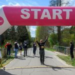 Osoby przechodzą przez kolorowy, różowo-biały nadmuchiwany łuk z napisem START. W tle widać drzewa pod błękitnym niebem. Teren wyścigu jest asfaltowy, otoczony zielenią.