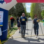 Grupa ludzi z numerami startowymi kończy marsz nordic walking. Pod nadmuchanym łukiem widać zieleń i słoneczne niebo. W tle las i wąska droga.