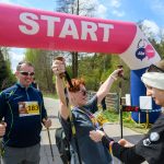 Start wyścigu nordic walking z różowym łukiem na tle zieleni. Kobieta z kijami unosi ręce w geście triumfu. Obok niej mężczyzna i wolontariuszka.