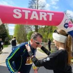 Mężczyzna odbiera medal od kobiety pod różowo-białym łukiem z napisem START. W tle zielone drzewa i niebo z chmurami. Na medal biegacza widnieje symbol i tekst.