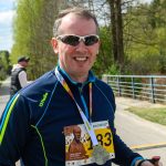 Na zdjęciu mężczyzna w okularach przeciwsłonecznych na tle zielonych drzew. Nosi niebieską kurtkę sportową i medal. W tle widać metalowe ogrodzenie i drogę.