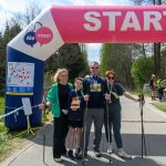Grupa ludzi stoi pod różowo-białym łukiem startowym na tle lasu. Mają medale i kijki do nordic walking. Pogoda jest słoneczna.