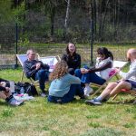 Grupa ludzi siedzi na trawie i leżakach, rozmawiając na zielonej polanie. W tle widać drzewa i ogrodzenie, sceneria jest spokojna i naturalna. Uczestnicy mają na sobie casualowe ubrania, a atmosfera jest relaksująca.