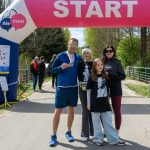 Grupa ludzi stoi pod różowym łukiem z napisem START. W tle drzewa i asfaltowa ścieżka. Jedna osoba trzyma medal.