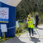 Zawodnik w zielonej czapce przechodzi przez linię mety pod niebiesko-czerwonym łukiem. Tło to drzewa i trawa. Obok stoi uśmiechnięta osoba.