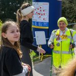 Zdjęcie przedstawia starszą osobę z kijkami do nordic walking w żółtej kamizelce i kapeluszu, rozmawiającą z dwiema dziewczynami. Tło to zielone krzewy i niebieski baner z mapą. Obok stoi dziewczyna w cz