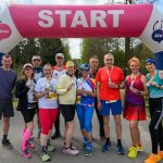 Grupa biegaczy z medalami stoi pod różowo-białym łukiem z napisem START. W tle drzewa i niebo. Uczestnicy są uśmiechnięci.