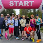 Grupa osób w kolorowych strojach sportowych stoi pod różowym łukiem START. Tło z zielonymi drzewami. Wszyscy trzymają medale.