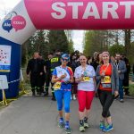 Trzy kobiety z medalami stoją przy różowej bramie startowej. Tło z zielonymi drzewami i ludźmi w strojach sportowych. Obok znajduje się zegar z czasem.