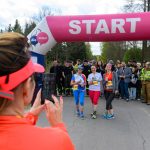 Trzy kobiety na starcie biegu, pod różowym banerem. W tle uczestnicy i strażacy. Kobieta w pomarańczowej bluzie robi zdjęcie.