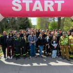Grupa ludzi stoi pod różowym balonem z napisem START. Tło z drzewami i niebieskim niebem. Uczestnicy w uniformach, niektórzy w kamizelkach odblaskowych.