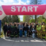 Grupa ludzi w mundurach stoi przed różowo-błękitnym napisem „START”. Tło stanowią drzewa i niebo. Atmosfera jest wiosenna i słoneczna.