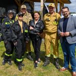 Grupa ludzi w mundurach stoi na trawie przed drewnianym budynkiem. Ubrania w odcieniach czerni i khaki, są napisy STRAŻ. W tle niebo z chmurami i zaparkowane samochody.