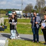 Na zdjęciu jest kilka osób na świeżym powietrzu. Mężczyzna przemawia do mikrofonu, a kobieta trzyma dyplom. Tło to zielona trawa, drzewa i samochody.