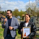 Na zdjęciu jest mężczyzna z mikrofonem i kobieta z ramką. Tło to zielona trawa i niebo z chmurami. Kobieta trzyma dyplom z podziękowaniem.