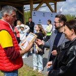 Mężczyzna w czerwonej kamizelce trzyma dyplom, otoczony grupą ludzi na zewnątrz. Tło: drewniana altana, zielona trawa. Słoneczny dzień z niebem pokrytym chmurami.