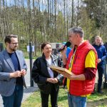 Na zdjęciu widać grupę ludzi na zewnątrz, przy mikrofonie. Tło stanowią drzewa i ogrodzenie, kolory są jasne i wiosenne. Osoby są uśmiechnięte, a atmosfera jest pozytywna.