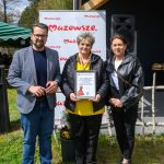 Trzy osoby stoją na trawie przed banerem z napisem Mazowsze. Jedna z nich trzyma certyfikat. Niebo jest błękitne i bezchmurne.