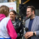 Trzy osoby rozmawiają na zewnątrz. Kobieta w różowej bluzie z nadrukiem Ale czas!. Mężczyzna trzyma mikrofon; tło jest zielone.