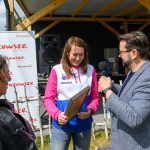 Na obrazie są trzy osoby na zewnątrz. Kobieta w biało-różowej bluzie trzyma certyfikat, rozmawiając z mężczyzną z mikrofonem. W tle jest baner Mazowsze i drewniana konstrukcja.