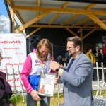 Trzy osoby stoją na zewnątrz, mężczyzna z mikrofonem gratuluje kobiecie. Kobieta trzyma dyplom, w tle baner Mazowsze. Pogoda jest słoneczna.