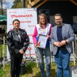 Trzy osoby stoją na tle tablicy z napisem Mazowsze, serce Polski. Osoba w środku trzyma dyplom. Scena znajduje się na zewnątrz, w słoneczny dzień.