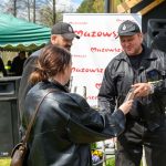 Na zdjęciu troje ludzi, w tym dwóch w mundurach, stoi na trawie. W tle zielony namiot i biały baner Mazowsze. Osoba wręcza coś drugiej.