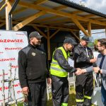 Grupa mężczyzn w czarnych uniformach uczestniczy w wydarzeniu na świeżym powietrzu. Jeden z nich w kamizelce odblaskowej podaje rękę mężczyźnie z mikrofonem. Tło to drewniana konstrukcja i baner z napisem Mazowsze.