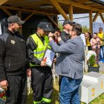 Na zdjęciu widać ceremonię wręczenia nagród na zewnątrz. Osoba w kamizelce odbiera medal, stojąc przed innymi ludźmi. W tle widoczne są tła z logotypem Mazowsze i podium.