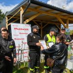 Grupa ludzi, w tym strażacy, na tle budynku pod drewnianą wiatą i plakatem Mazowsze. Przyznawanie dyplomów, kolorowe stroje. Słoneczny dzień z chmurami.