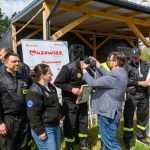 Grupa strażaków otrzymuje nagrody na tle drewnianej altanki. W prawym górnym rogu widać baner Mazowsze. Strażacy ubrani są w czarne uniformy z żółtymi elementami.