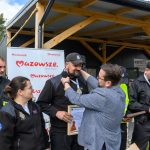 Grupa strażaków w czarnych mundurach stoi przed drewnianą altaną. Jedna osoba odbiera medal, w tle baner Mazowsze. Na dworze jest słonecznie, wokół widać drzewa i budynek.