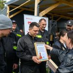 Grupa strażaków w czarnych mundurach odbiera dyplom. Tło to drewniana konstrukcja i baner z napisem Mazowsze. Atmosfera jest uroczysta, a ludzie uśmiechnięci.