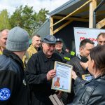 Grupa osób w mundurach otrzymuje dyplom, tło zielone drzewo i namiot. Mężczyzna trzyma ramkę, obok mikrofon. Pogodne niebo.