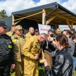 Grupa strażaków w czarnych i beżowych mundurach odbiera nagrody na tle budynku. Tło to zielona trawa i niebieskie niebo. W tle widoczny jest baner z napisem Mazowsze.