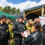Grupa strażaków w czarnych i beżowych mundurach odbiera certyfikaty. Tło to zielony namiot i budynek, z drzewami i niebem. Słoneczny dzień, atmosfera uroczysta.