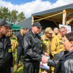 Grupa strażaków w czarnych i żółtych mundurach stoi na zewnątrz, przy namiocie. Jeden z nich podaje dokument kobiecie w skórzanej kurtce. Tło stanowi budynek z drewna i zielony namiot.