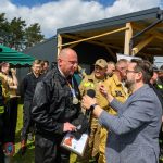 Grupa ludzi w różnych mundurach stoi na trawie obok budynku z drewnianym dachem. Jeden mężczyzna trzyma mikrofon. W tle zielone drzewa i fragment nieba pokrytego chmurami.