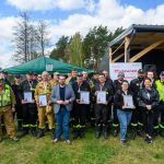 Grupa strażaków w mundurach stoi na trawie, trzymając dyplomy. W tle zielony namiot i drewniany budynek. Niebo jest niebieskie z chmurami.
