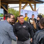 Grupa ludzi przed budynkiem z drewnianym zadaszeniem. Zielone tło z drzewami, plakat z napisem Gmina Dobre. Mężczyźni podają sobie ręce.